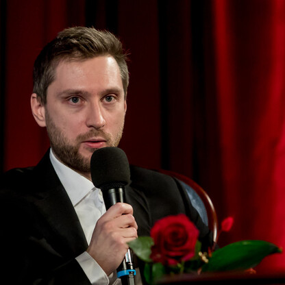 Mężczyzna w czarnej kamizelce i białej koszuli, trzymający mikrofon, siedzi przed czerwoną zasłoną, uczestnicząc w panelu dyskusyjnym. (wygenerowano przez AI)