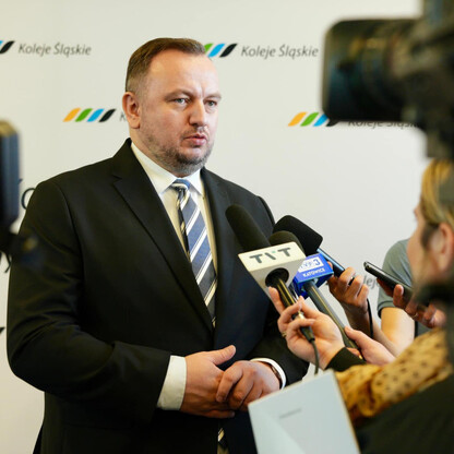 Na zdjęciu: Mężczyzna w garniturze podczas konferencji prasowej, otoczony mikrofonami i kamerami, z logo 