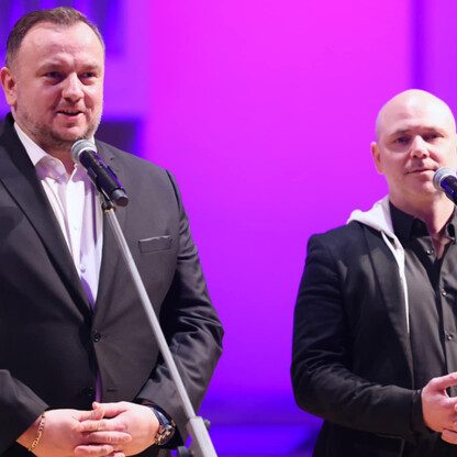 Mężczyzna w czarnej这套西装站在舞台上，手持麦克风，似乎在进行演讲或表演，背景为鲜艳的紫色，可能是在“Muzyczna Tradycyja”活动中。 (wygenerowano przez AI)