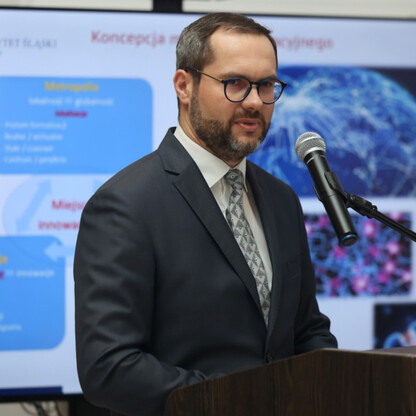 Mężczyzna w garniturze przemawia na podium, tło: prezentacja 