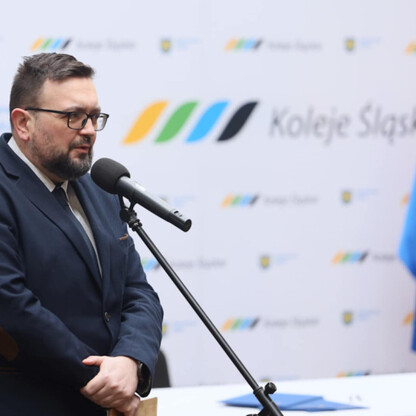 Mężczyzna w ciemnym garniturze i okularach przemawia do mikrofonu na konferencji prasowej Kolei Śląskich. (wygenerowano przez AI)