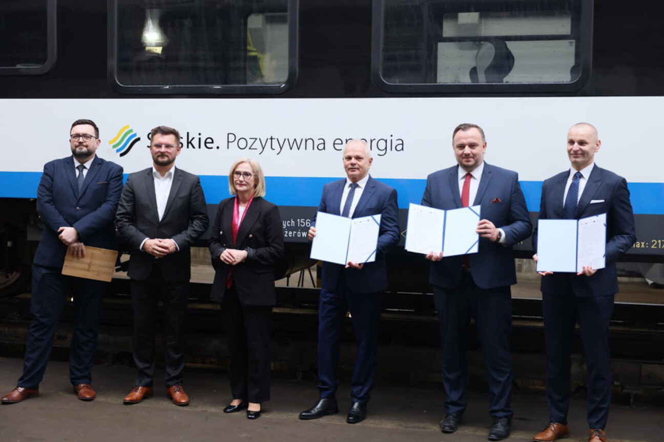 Mężczyzna w garniturze przemawia na podium z mikrofonem, przedstawiając umowę o dostawę 26 elektrycznych zespołów trakcyjnych dla Kolei Śląskich. (wygenerowano przez AI)