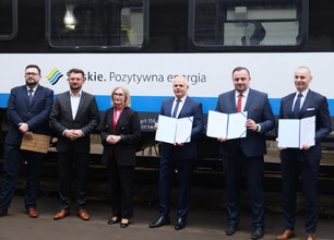 Mężczyzna w garniturze przemawia na podium z mikrofonem, przedstawiając umowę o dostawę 26 elektrycznych zespołów trakcyjnych dla Kolei Śląskich. (wygenerowano przez AI)