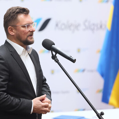 Mężczyzna w garniturze przemawia na podium z mikrofonem, przedstawiając nowe elektryczne zespoły Kolei Śląskich. (wygenerowano przez AI)