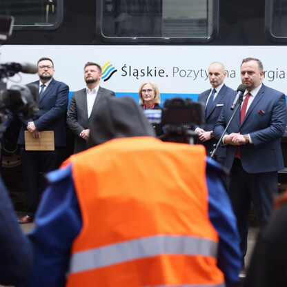 Grupa ludzi z mikrofonami i kamery przygotowuje się do wydarzenia związanych z dostawą 26 elektrycznych zespołów trakcyjnych do Kolei Śląskich. (wygenerowano przez AI)