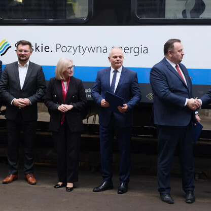 Grupa oficjalnych osób w formalnym stroju podpisuje dokumenty przed pociągiem z napisem 
