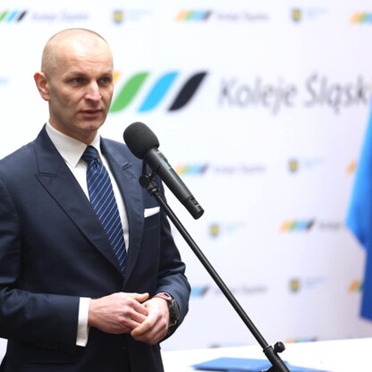 Mężczyzna w garniturze przemawia na podium z mikrofonem, przedstawiając umowę o dostawę 26 elektrycznych zespołów trakcyjnych dla Kolei Śląskich. (wygenerowano przez AI)