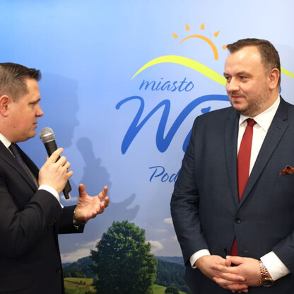 Dwóch mężczyzn w strojach formalnych, jeden trzymający mikrofon, przed logo 