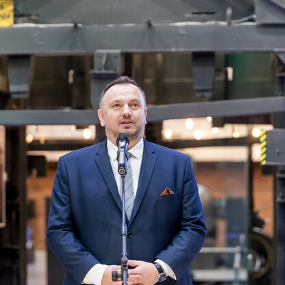 Mężczyzna w niebieskim garniturze przemawia na podium w dawnej kopalni, podkreślając znaczenie dziedzictwa przemysłowego. (wygenerowano przez AI)
