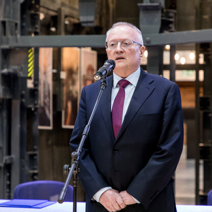 Mężczyzna w ciemnym garniturze i czerwonym krawacie stoi na podium z mikrofonem, przemawia w Kopalni Ignacy. (wygenerowano przez AI)