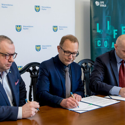 Trzech mężczyzn w strojach formalnych siedzi przy stole, podpisywani dokumenty. Regionalna inicjatywa województwa Śląskiego do adaptacji do zmian klimatu. (wygenerowano przez AI)