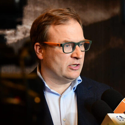 Mężczyzna w garniturze i okularach przemawia do mikrofonu z logo 
