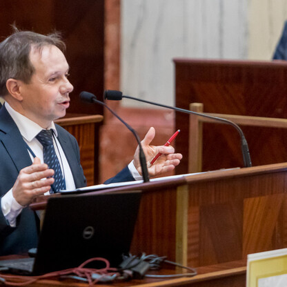 Na zdjęciu: Mężczyzna w garniturze uczestniczący w spotkaniu geodezyjnym, gestykuluje podczas dyskusji. (wygenerowano przez AI)