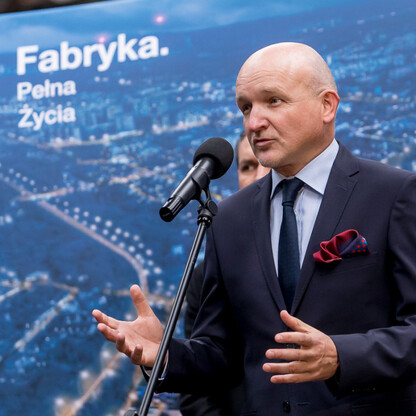 Mężczyzna w ciemnym garniturze przemawia na podium z mikrofonem, przedstawiając projekt rewitalizacji fabryki Defum w Dąbrowie Górniczej. (wygenerowano przez AI)
