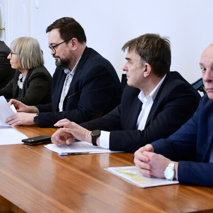 Grupa pięciu osób w strojach formalnych siedzi przy długim drewnianym stole w sali konferencyjnej, dyskutując o reaktywacji linii kolejowej Gliwice-Rybnik. (wygenerowano przez AI)