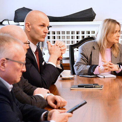 Grupa ludzi w formalnym ubiorze siedzi wokół długiego drewnianego stołu w Gliwickim Sejmiku Śląskim, omawiając plany dotyczące odbudowy linii kolejowej Gliwice-Rybnik. (wygenerowano przez AI)