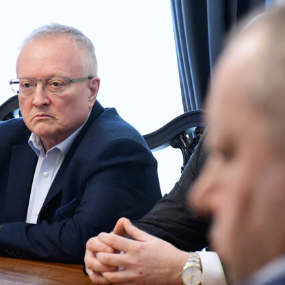 Mężczyzna w ciemnym garniturze i okularach siedzi przy stole, uczestnicząc w serio traktowanym spotkaniu. (wygenerowano przez AI)