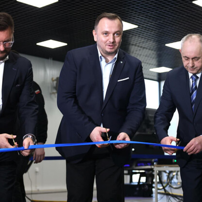 Trzy mężczyźni w garniturach uczestniczą w uroczystości otwarcia Krajowego Centrum Bezpieczeństwa Ruchu Drogowego w Katowicach. Przygotowują się do przecięcia niebieskiego sznurka. (wygenerowano przez AI)