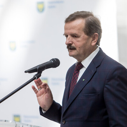 Mężczyzna w ciemnym garniturze przemawia do mikrofonu, gestykulując, podczas symbolicznego przekazania funduszy na drogi dojazdowe do gruntów rolnych w południowym Śląsku. (wygenerowano przez AI)